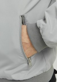 Hellgraue Bomberjacke mit einer Reißverschlusstasche und gerippten Bündchen. Sie verfügt über eine glatte Stoffstruktur und metallene Reißverschlussbeschläge.