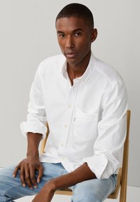 Homme aux cheveux courts portant une chemise blanche à boutons et un jean bleu clair, assis sur une chaise en bois devant un fond uni.