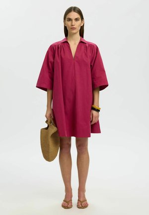 Vrouw in een losse, knielange magentakleurige jurk, met een bruine gevlochten tas, armbanden om één pols en beige sandalen.