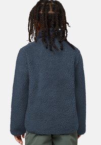 Enfant avec des cheveux torsadés de longueur moyenne, portant une veste en polaire texturée bleu marine et un pantalon vert, vu de dos.