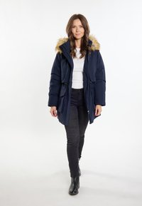 Marineblå polstret parka-jakke med hætte i kunstpels, lynlås foran, snoretræk i taljen og sidelommer. Båret over en hvid top og sorte bukser.