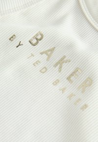 Nahaufnahme eines weißen Rippenstoffs mit diagonal aufgedrucktem goldfarbenem metallischem Schriftzug „BAKER BY TED BAKER“.
