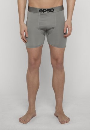 Hombre con bóxers PSD grises de corte ajustado, de pie descalzo frente a un fondo gris liso, con las manos relajadas a los lados.
