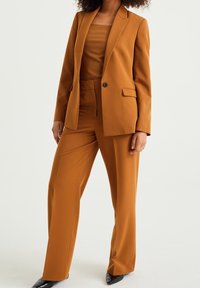 Orangebrun kostym med en skräddarsydd blazer med en knapp och fickdetaljer, kombinerad med vida byxor och en ribbad topp.