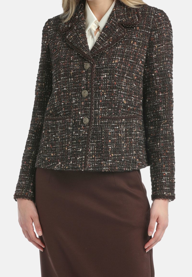 Femme portant un blazer en tweed marron foncé avec des taches multicolores, trois boutons décoratifs et une jupe marron sur un fond clair.