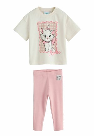 Young girl in white shirt with pink cat graphic and pink leggings, smiling and posing with one foot raised against a light background.