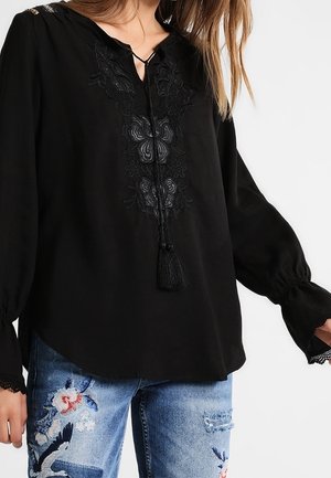 Mujer con blusa negra de manga larga con bordado floral y lazos con borlas, combinada con vaqueros azules con parches florales y de pájaros.