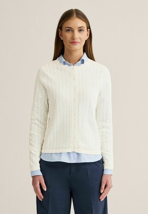 Kvinde iført hvid tekstureret knappet cardigan over en lyseblå skjorte og mørkeblå bukser, stående foran en ensfarvet beige baggrund.