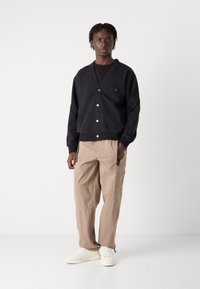 Black cardigan with a V-neck, white buttons, and patterned accents; paired with light beige cargo pants and white sneakers.