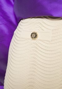 Tissu tricoté de couleur crème avec une texture ondulée, orné d'un bouton fleur décoratif en métal doré. Tissu violet en arrière-plan.