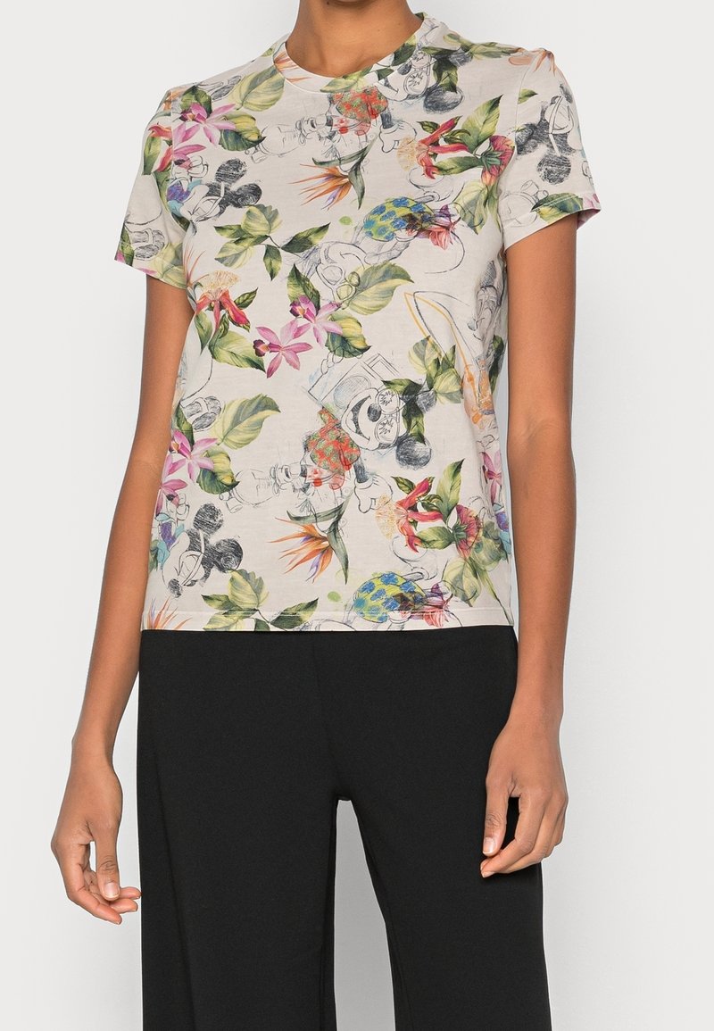 Kvinna som bär en kortärmad beige t-shirt med färgglada blommönster och tecknade figurer, kombinerat med svarta byxor.