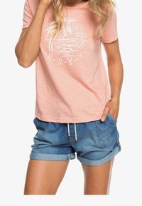 T-shirt rose clair avec un motif de palmier blanc et le texte "Roxy". Associé à un short en denim retroussé avec taille à cordon et poches.