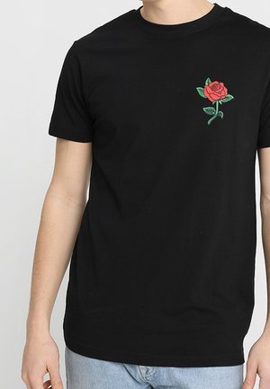 Černé tričko s krátkým rukávem, na levé straně hrudi vyšívaná malá červená růže se zelenými listy, nošené osobou ve světle modrých džínách.