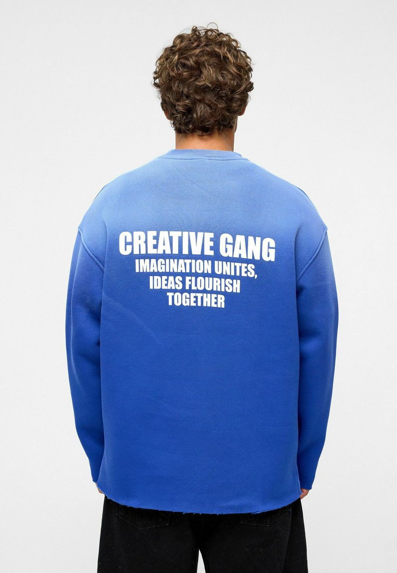 Blauwe sweatshirt met een iets oversized pasvorm, voorzien van een ruwe zoom en witte grafische tekst op de achterkant: "CREATIVE GANG IMAGINATION UNITES."
