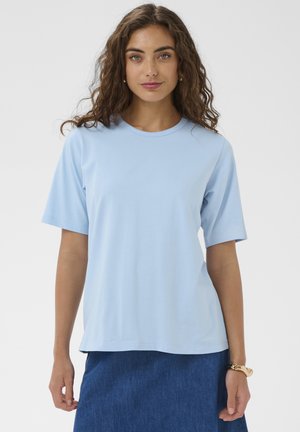 Femme aux cheveux bruns bouclés portant une chemise bleu clair à manches courtes et une jupe bleu foncé, debout devant un fond blanc uni.