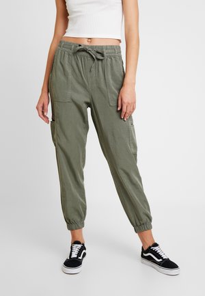 Kobieta w oliwkowo-zielonych spodniach typu jogger cargo z elastycznym paskiem i czarnych sneakersach, stojąca na jednolitym tle.