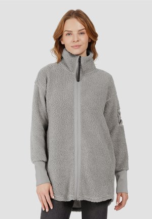 Woman wearing a light gray, high-collar textured fleece jacket with long sleeves and a front zipper, standing against a plain background.