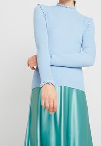 Himmelblaues, geripptes Langarmoberteil mit Rüschen am Hals und an den Ärmelbündchen, kombiniert mit einem glänzenden petrolfarbenen Rock.