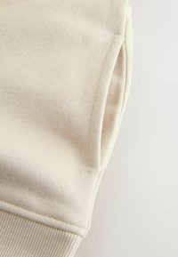 Tejido de sudadera color beige claro con detalles de dobladillo y puños acanalados. Textura suave, con un práctico bolsillo lateral. Diseño minimalista.