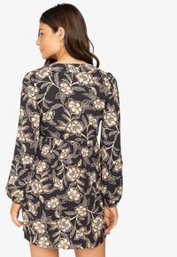 Vestido negro con flores y mangas largas, que presenta una cintura ajustada, patrones florales en beige claro y dorado, y un acabado de tela texturizada.