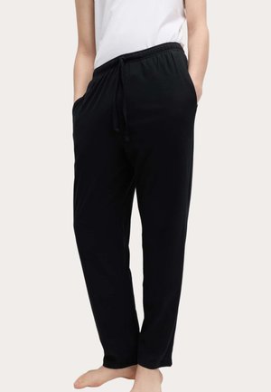 Person trägt lockere schwarze Kordelzughose mit den Händen in den Taschen, kombiniert mit einem weißen Kurzarmoberteil, barfuß vor einfachem Hintergrund.