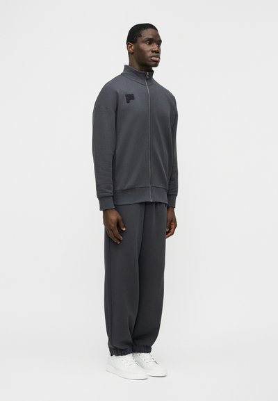 Homme debout portant une veste de survêtement zippée gris foncé et un pantalon assorti avec des baskets blanches sur un fond clair uni.