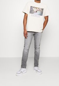 Camiseta de algodón color crema con un gráfico de "El Exorcista" en letras moradas, combinada con vaqueros gris ajustados y zapatillas blancas con detalles en gris.