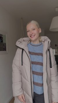 Lysebeige dunjakke med hætte, sorte lynlåse, over en blå trøje med brune og blå striber. Ensfarvet baggrund med minimal dekoration.