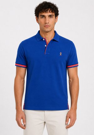 Homme aux cheveux bouclés et foncés portant un polo bleu à manches courtes avec une bordure rouge et un pantalon beige, face à l'avant sur un fond uni.
