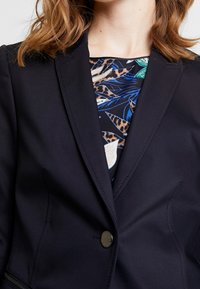 Granatowa marynarka z pojedynczym guzikiem, strukturalnymi klapami i gładką teksturą. Nosi się ją na wzorzystej bluzce z niebieskimi liśćmi i zwierzęcym nadrukiem.