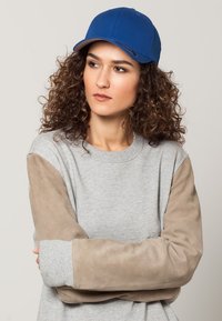 Cappellino da baseball blu con visiera curvata, abbinato a una felpa grigia chiara con maniche in suede marrone. Dettagli del tessuto e del design chiaramente visibili.