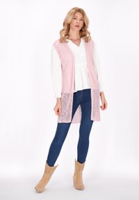 Lichtroze gebreide vest met open voorkant, gedragen over een witte blouse en blauwe spijkerbroek. Gecombineerd met beige cowboylaarzen, met een textuur en een strakke pasvorm.