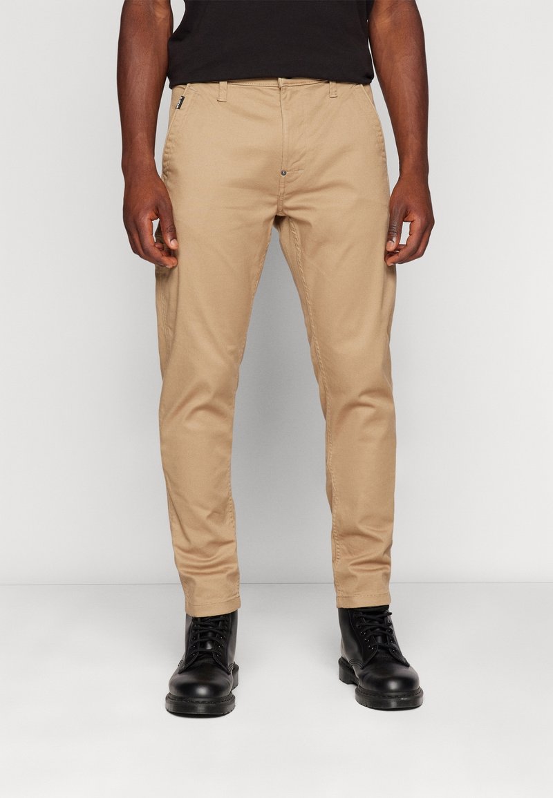 Homme portant un pantalon beige ajusté, une chemise noire et des bottes noires à lacets, debout devant un fond clair uni.