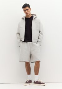 Tânăr purtând hanorac gri cu glugă și pantaloni scurți asortați, tricou negru, șosete albe și adidași mov, stând sprijinit de un perete alb cu mâinile în buzunare.