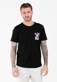 Hombre con los brazos tatuados, vistiendo una camiseta negra con un diseño floral en el pecho y pantalones blancos, de pie contra un fondo blanco.