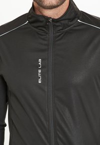 Zwarte sportjack met een hoge kraag, rits aan de voorkant en reflecterende accenten. Heeft een gladde, rekbare stof met een subtiele glans.