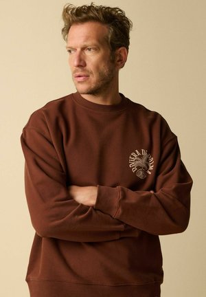 Homme aux cheveux bouclés portant un sweat-shirt marron avec un petit motif de palmier et un texte sur la poitrine gauche, bras croisés, regardant vers la gauche.