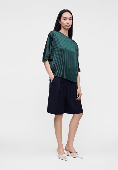 Femme aux longs cheveux foncés portant un haut plissé ample vert foncé, un short noir longueur genou et des talons hauts pointus rose clair.