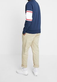 Man med marinblå tröja med vita och röda ränder på ärmarna, beiga uppvikta byxor, vita strumpor och vita sneakers, stående med händerna i fickorna.