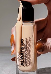 Flacon de fond de teint Smashbox Always On avec un fond transparent, une pompe noire et un liquide tan s'écoulant de la buse ; texte en noir sur le flacon.