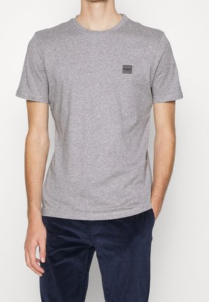 Mann trägt ein schlichtes graues kurzärmliges T-Shirt mit einem kleinen "BOSS"-Logo-Patch auf der Brust, kombiniert mit dunkelblauen Cordhose.