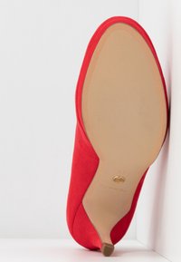 Une chaussure à talon haute en suède rouge reposant, avec la semelle vers le haut, contre un mur blanc de coin, montrant une semelle beige lisse et un petit embout de talon.