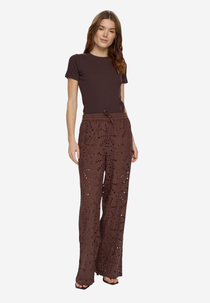 Femme portant un haut marron foncé ajusté à manches courtes et un pantalon large et ample en dentelle ajourée marron avec une taille à cordon.
