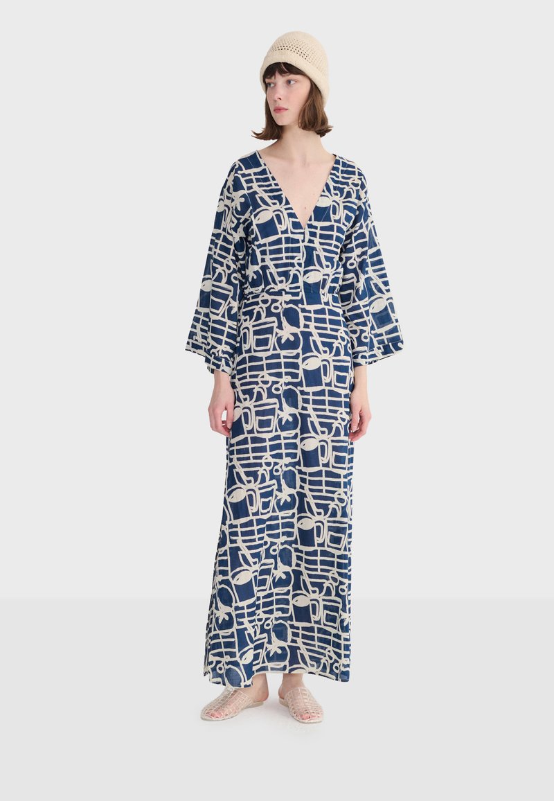 Kobieta w długiej sukience w granatowo-biały wzór z szerokimi rękawami, beżową dzianinową czapką i plecionymi wsuwanymi butami, stojąca na jednolitym tle.