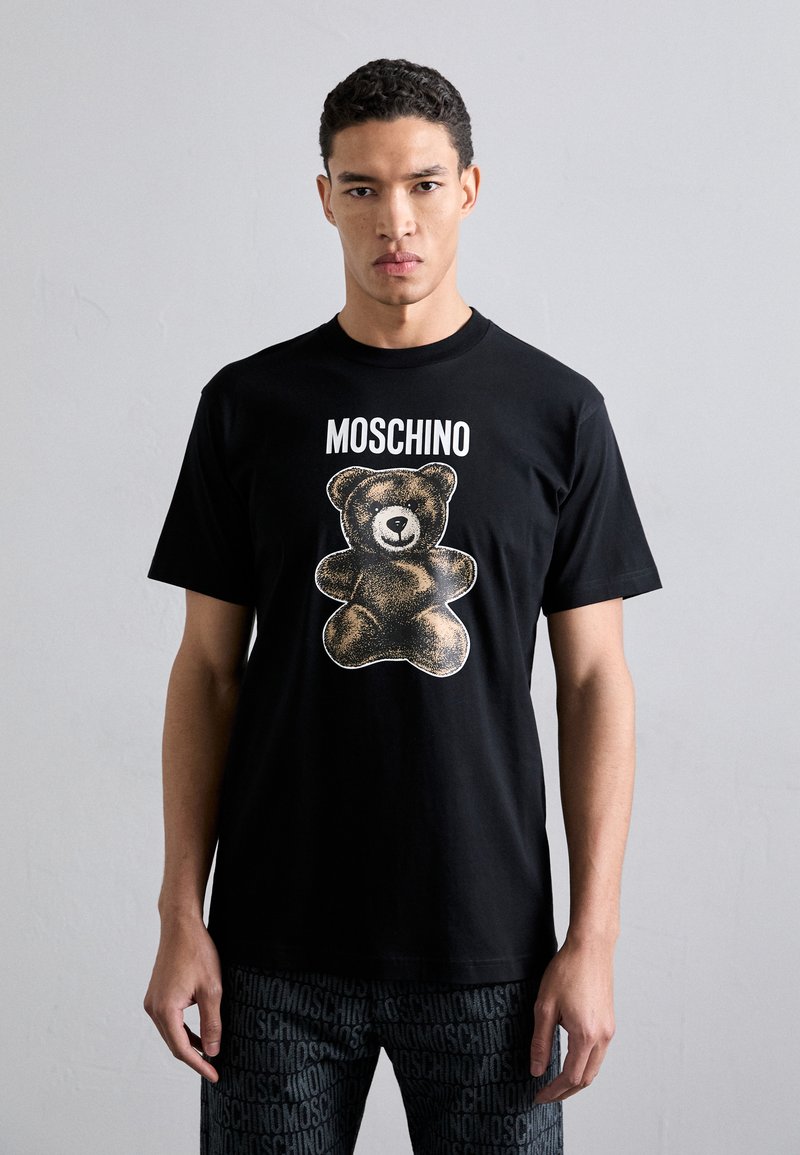 T-shirt en coton noir avec un grand motif d'ours en or et blanc, avec le texte "MOSCHINO" au-dessus de l'ours. Col rond, manches courtes.