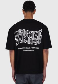 Zwarte katoenen t-shirt met grote witte tekst op de achterkant. Ontwerp bevat "DROPSIZE" en "DROPSIZE CLUB / EST. 2020." Korte mouwen.