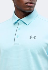 Άνδρας που φοράει ανοιχτό μπλε πόλο Under Armour με τρία κουμπιά και λογότυπο στο στήθος, θέα εστιασμένη στον γιακά και στο πάνω μέρος του κορμού.