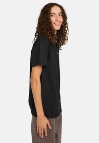 T-shirt nera in cotone con maniche corte, taglio dritto e una texture morbida. Indossata con pantaloni marroni. Design semplice, senza pattern visibili.