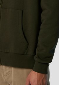 Felpa con zip di colore verde scuro, dotata di tasca a marsupio, polsini e orlo a coste, realizzata in materiale morbido, mostrata da vicino sul braccio e sul lato.