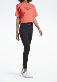 Červené zkrácené tričko s černým logem Reebok, v kombinaci s černými legínami a bílými teniskami. Elegantní, přiléhavý design s hladkými texturami.